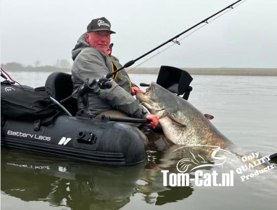 Een persoon in een grijze jas en een pet houdt een grote vis vast terwijl hij in een opblaasbare kajak op het water zit.