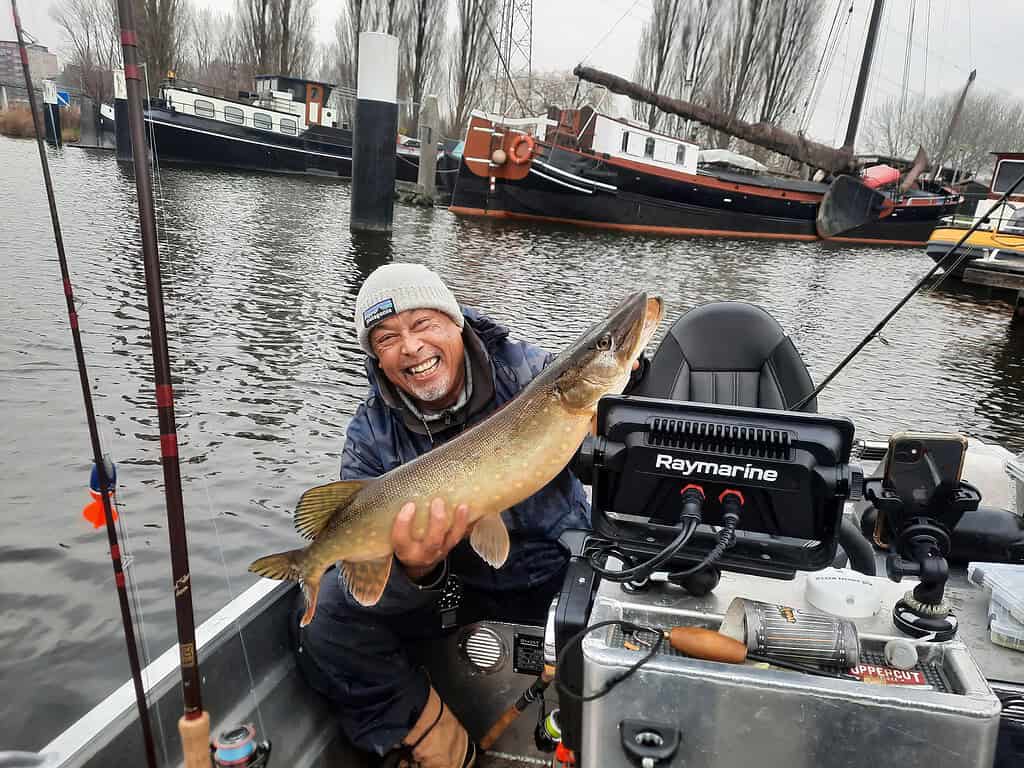 Een persoon die lacht terwijl hij een grote vis vasthoudt op een boot, met indrukwekkende vangsten. Vishengels en navigatieapparatuur zijn zichtbaar.