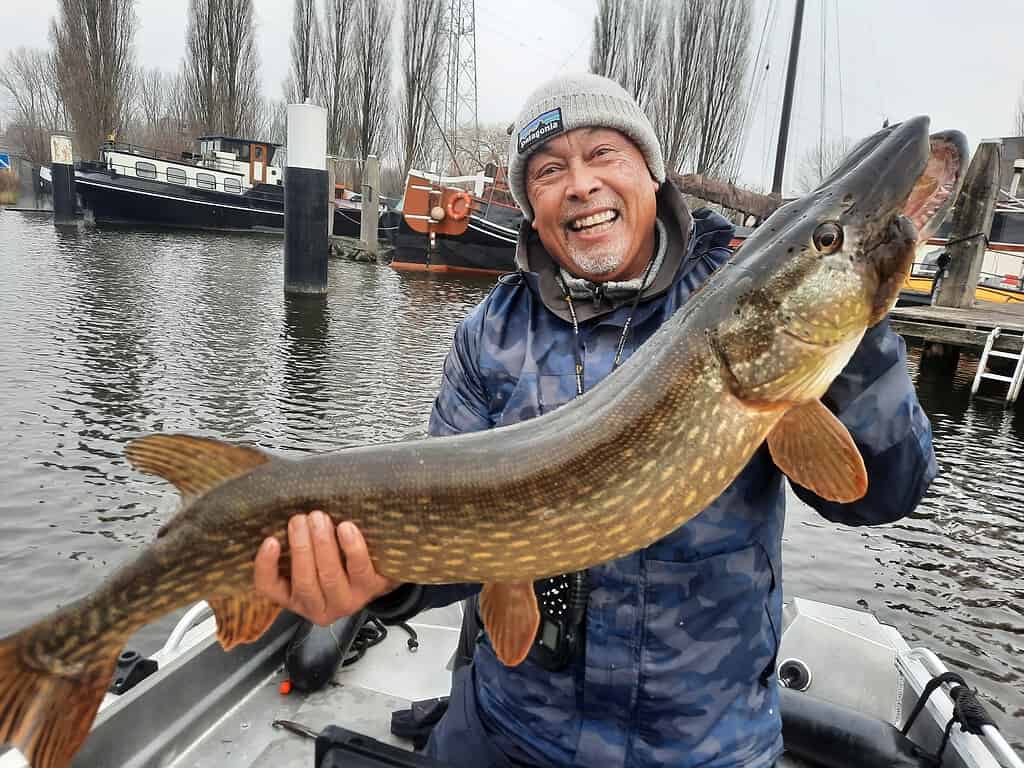 Een lachende persoon houdt trots een grote vis vast op een laars, met water en andere boten op de achtergrond, die de opwinding van Vangsten vastleggen.