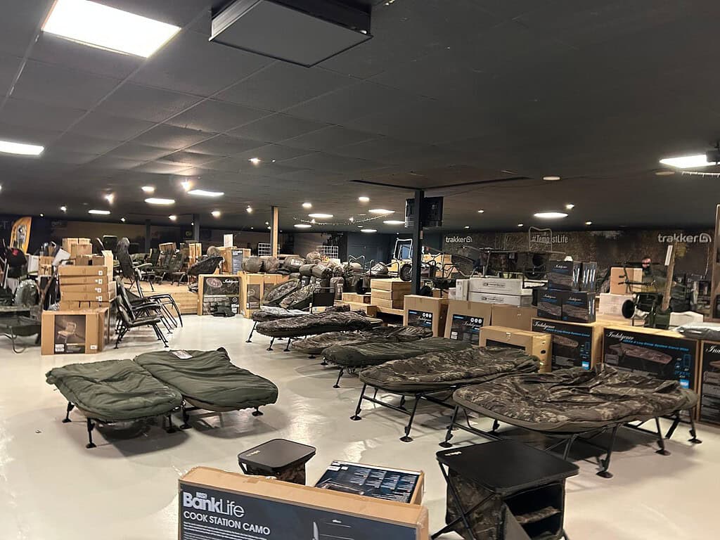 Het interieur van de winkel toont een scala aan kampeer- en outdooruitrusting, waaronder stoelen en tafels met camouflagepatroon die netjes in rijen zijn gerangschikt. Ontdek de nieuwste Hareco Karper-collecties of bezoek de Open Dag voor exclusieve deals.