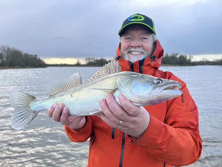 Een man in een oranje jas en een groene hoed glimlacht terwijl hij een grote vis vasthoudt aan de rand van het water.