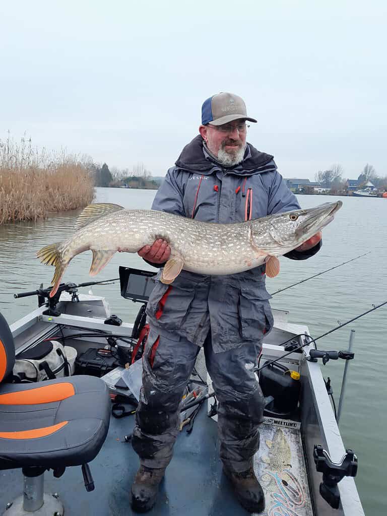 Een man staat trots op een boot en toont zijn vangsten, een grote vis die hij ving in de onstuimig wateren van het jaar. Riet en glinsterend water creëren een pittoresk decor.