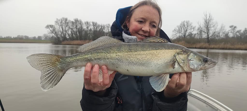 Een persoon die trots een grote vangsten vasthoudt op de boot, met een mistig meer dat het tafereel vormt te midden van een onstuimig week. Bomen staan majestueus op de achtergrond.