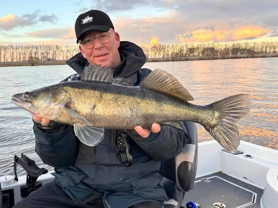 Een man met een zwarte pet houdt een grote vis vast op een boot, met een kalm meer en een rij bomen op de achtergrond bij zonsondergang.