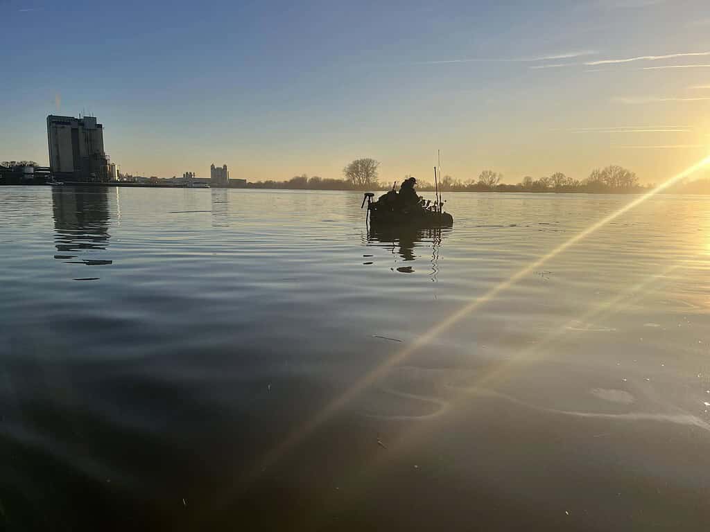 Een persoon in een klein bootje op een kalm meer bij zonsondergang, ervaart in alle rust het gevoel van rust, terwijl in de verte gebouwen zichtbaar zijn.