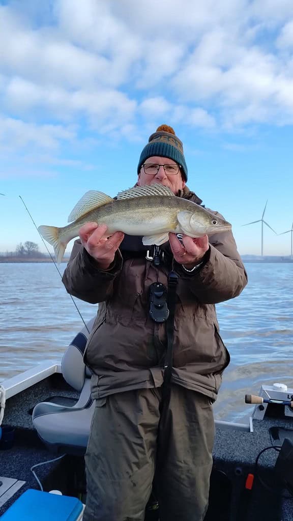 Tegen de achtergrond van windturbines en water staat een persoon trots op een boot, met zijn nieuwste vangst in zijn handen. Gekleed in een muts en winterjas, glimlachen ze bij de belofte van betere vangsten terwijl ze genieten van dit koude avontuur.