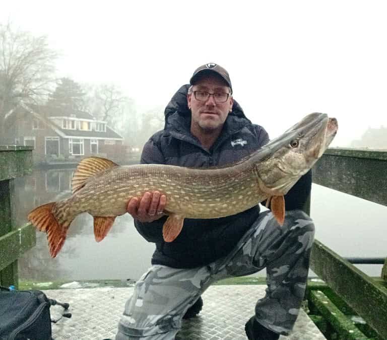 Man met een grote snoekvis op een houten brug bij een waterpartij, met mistige bomen en huizen op de achtergrond.