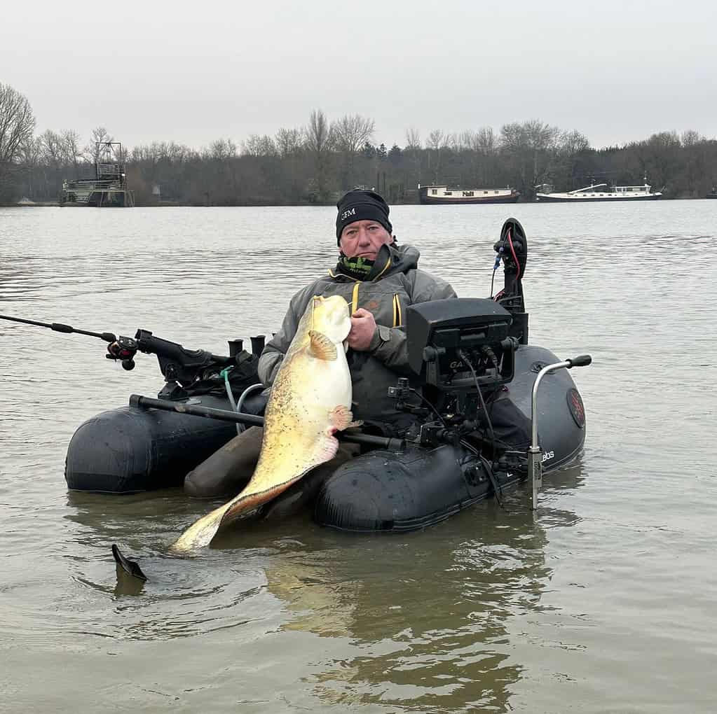 Persoon in een kleine opblaasbare vissersboot die een grote vis vasthoudt op een bewolkte dag, met bomen en een boot op de achtergrond.