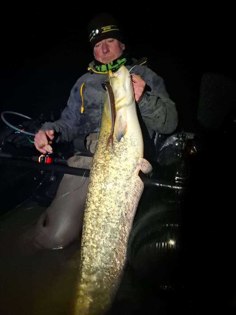 In de frisse nachtlucht zit een persoon, gekleed in warme winterkleding, triomfantelijk in zijn boot, nadat hij net gestopt is met vechten tegen een monsterlijke vis. De vangst schittert in het maanlicht en toont een zwaarbevochten overwinning.