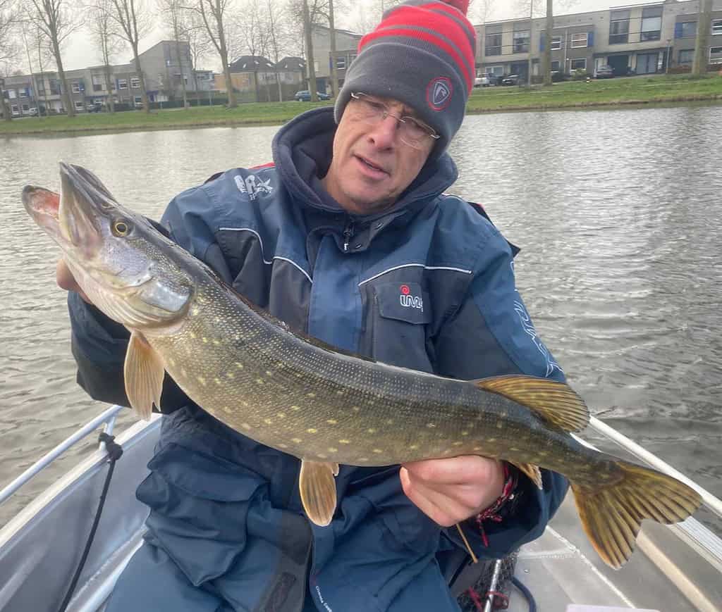 Een persoon in een bootje toont trots zijn vangsten, met een wintermuts en jas op tegen de koude lucht, met het kalme water en de schilderachtige huizen op de achtergrond.