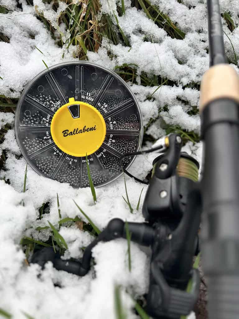 Visgerei, waaronder een haspel en een houder met het opschrift "Ballabeni", wordt op het met sneeuw bedekte gras gelegd, perfect voor een winters penvisavontuur.