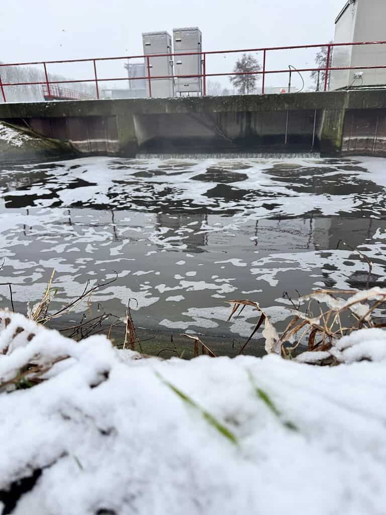 Met sneeuw bedekte grond op de voorgrond met een watermassa die schuim vertoont, perfect voor winterpenvissen. Op de achtergrond doemt een betonnen dam met metalen leuningen en industriële structuren groot op.