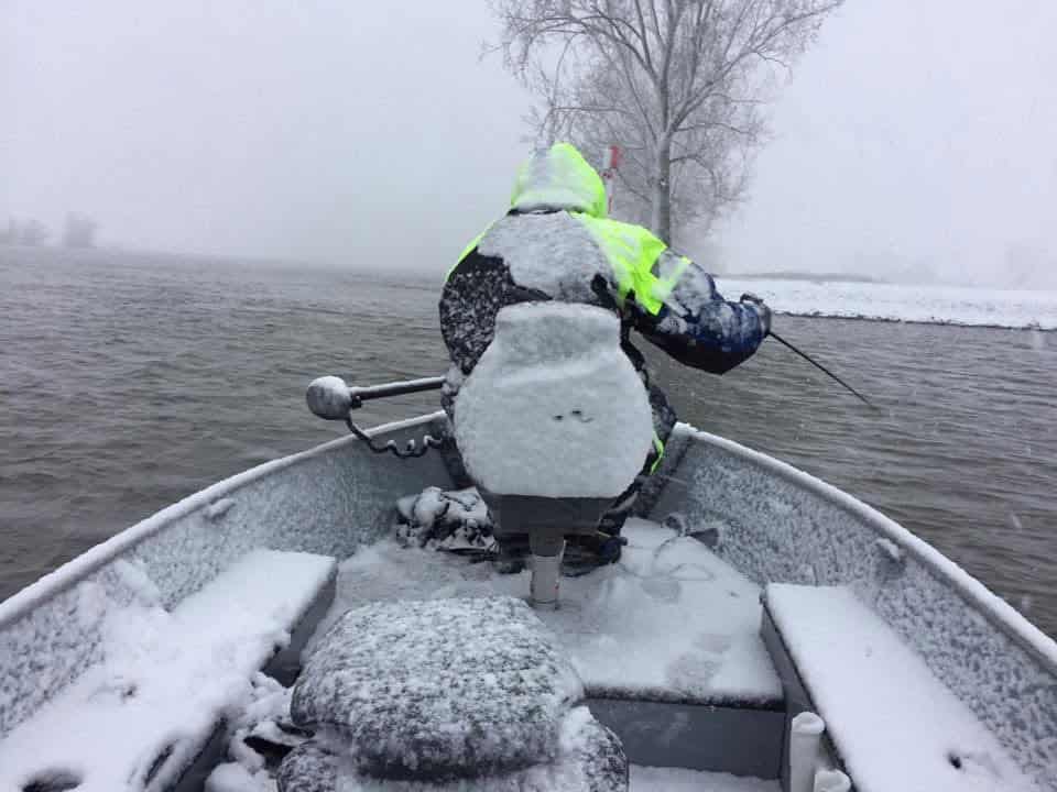 Een persoon in winterkleding is bezig met wintervissen op een met sneeuw bedekte boot, omringd door de rustige schoonheid van een besneeuwde rivier, geduldig hopend dat het gladde dakvis bijt.