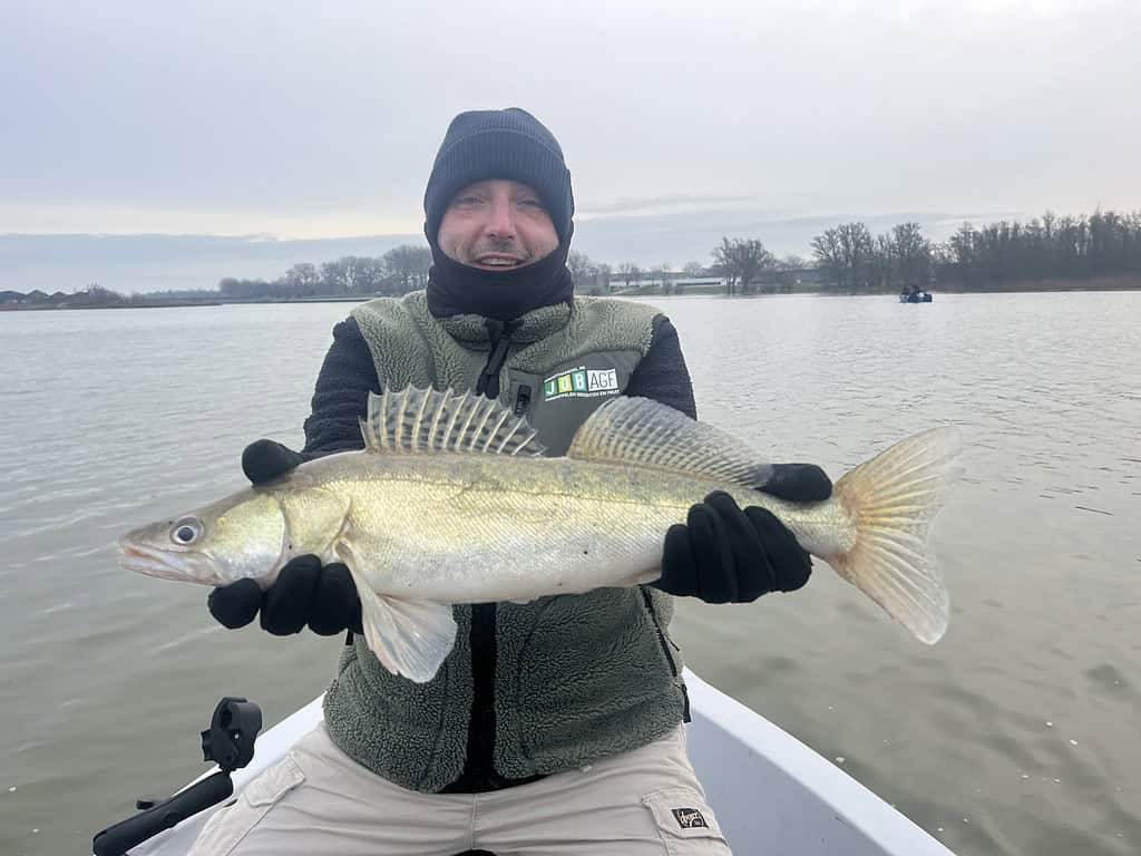 Een persoon is gehuld in een muts en handschoenen en geniet van de wintervisserij. Hij of zij houdt trots een roofvis vast en zit in een bootje op het rustige meer.