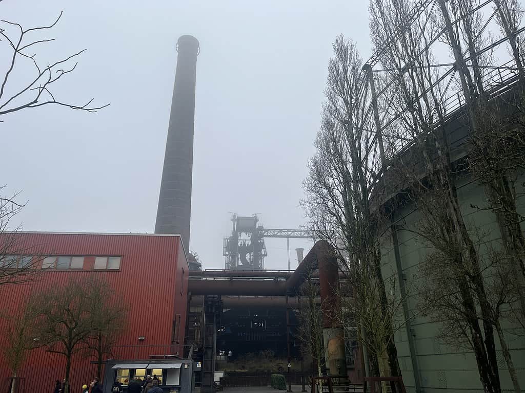 Het industrieterrein, dat doet denken aan de Angelmesse Duisburg, staat met zijn torenhoge schoorsteen, rode gebouw en grote tank te midden van bladloze bomen op een mistige dag.