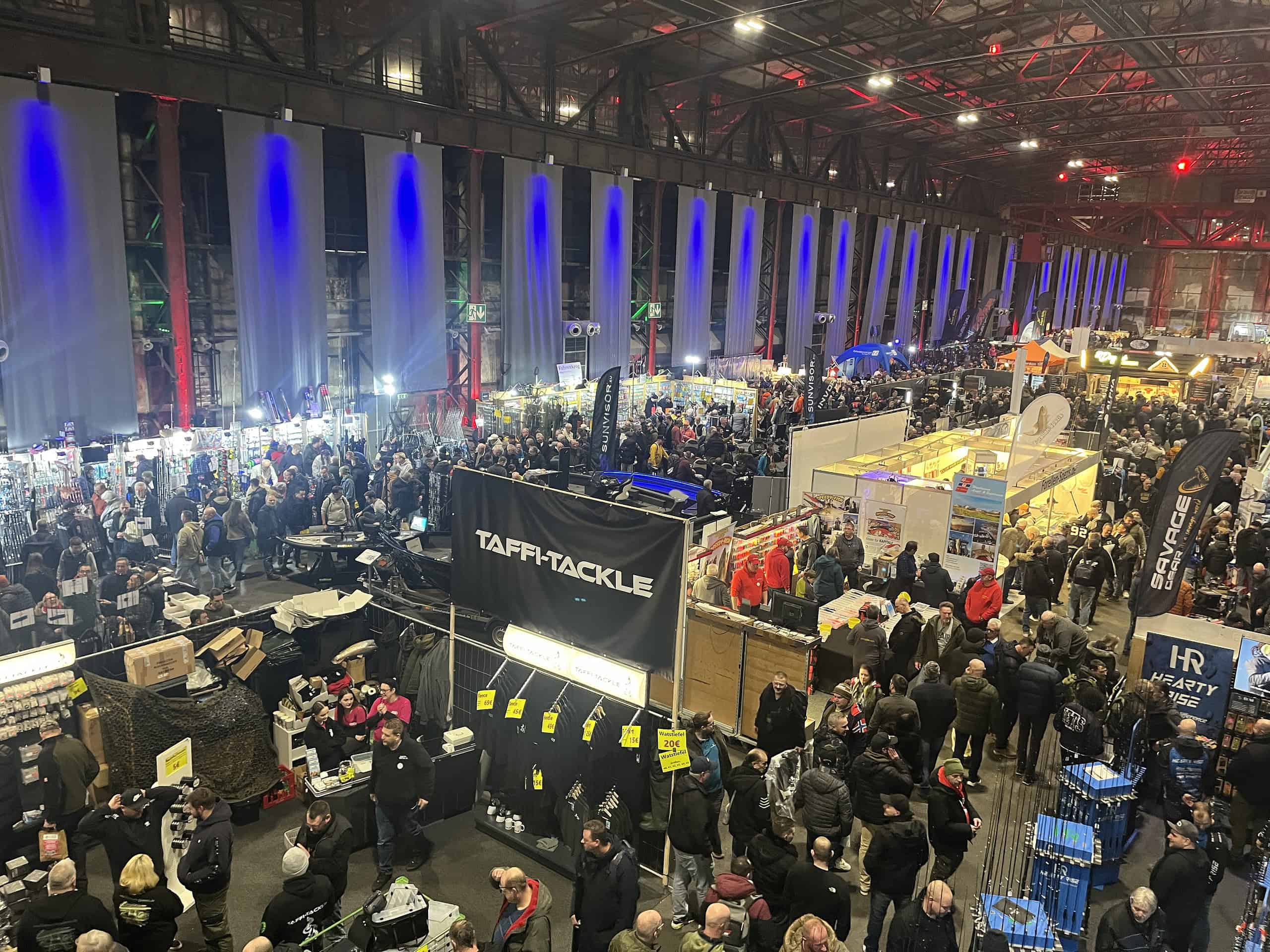 Een grote menigte verzamelt zich in het Angelmesse Duisburg congrescentrum, met verschillende stands die visgerei en accessoires onder felle verlichting tentoonstellen. Het is zo druk dat je "over de koppen" zou kunnen lopen, terwijl enthousiastelingen de nieuwste innovaties op het gebied van visgerei verkennen.