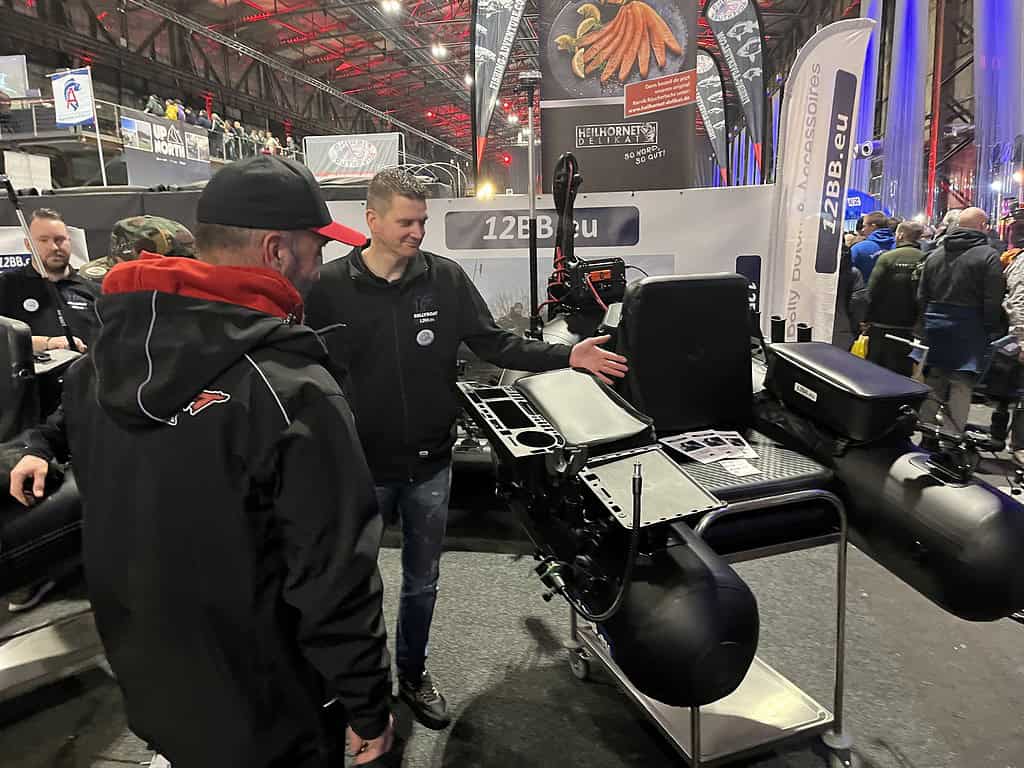 Op het bruisende Angelmesse Duisburg-evenement demonstreert een man apparatuur aan een ander persoon te midden van een zee van stands en banners. De sfeer is levendig, met bezoekers die praktisch "lopen over de koppen" in hun enthousiasme om de nieuwste visserijinnovaties te zien.