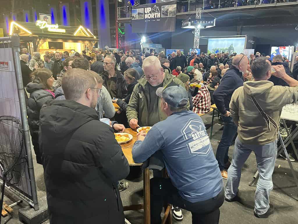 Bij Angelmesse Duisburg ontvouwt zich een bruisende bijeenkomst binnenshuis met mensen die rond tafels staan te kletsen en genieten van eten en drinken. Felle verlichting benadrukt de levendige banners op de achtergrond, wat bijdraagt aan de energieke sfeer.