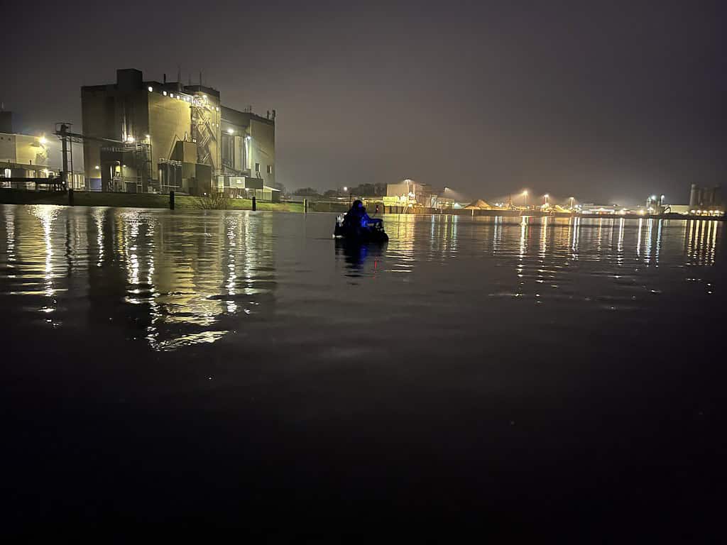 Een eenzame kanoër peddelt 's nachts op kalm water, vlak bij felverlichte industriële gebouwen en bruggen.