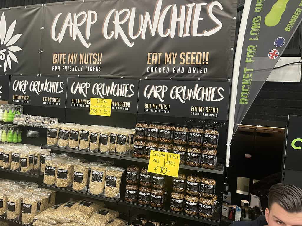 Stand met Carp Crunchies visvoer in verschillende verpakkingsformaten op schappen op de gezellige beurs. Borden benadrukken beursprijzen en productbeschrijvingen, waardoor het een must-see is op Carp Den Bosch 2025.