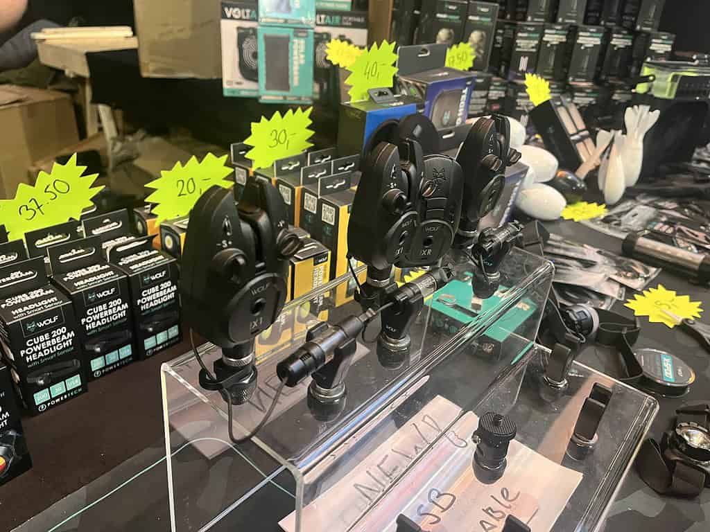 Er hangt een levendige sfeer in de winkel, waar allerlei elektronische apparaten met prijskaartjes op een tafel staan uitgestald. Het doet denken aan de drukke beurs op Carp Den Bosch, waar diverse gadgets op een transparante standaard zijn gemonteerd.