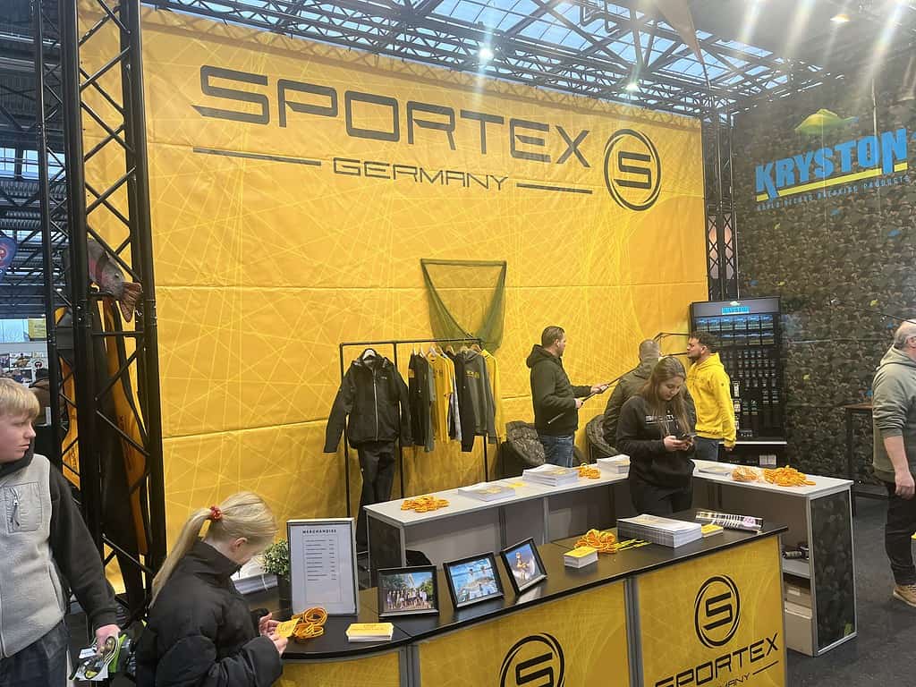 Op de stand van Sportex Germany kunnen bezoekers van de beurs van 2025 een scala aan visgerei en -kleding ontdekken. Onder de felle verlichting snuffelen en staan geïnteresseerden in Carp Den Bosch in de buurt, gretig om de nieuwste innovaties te ontdekken.