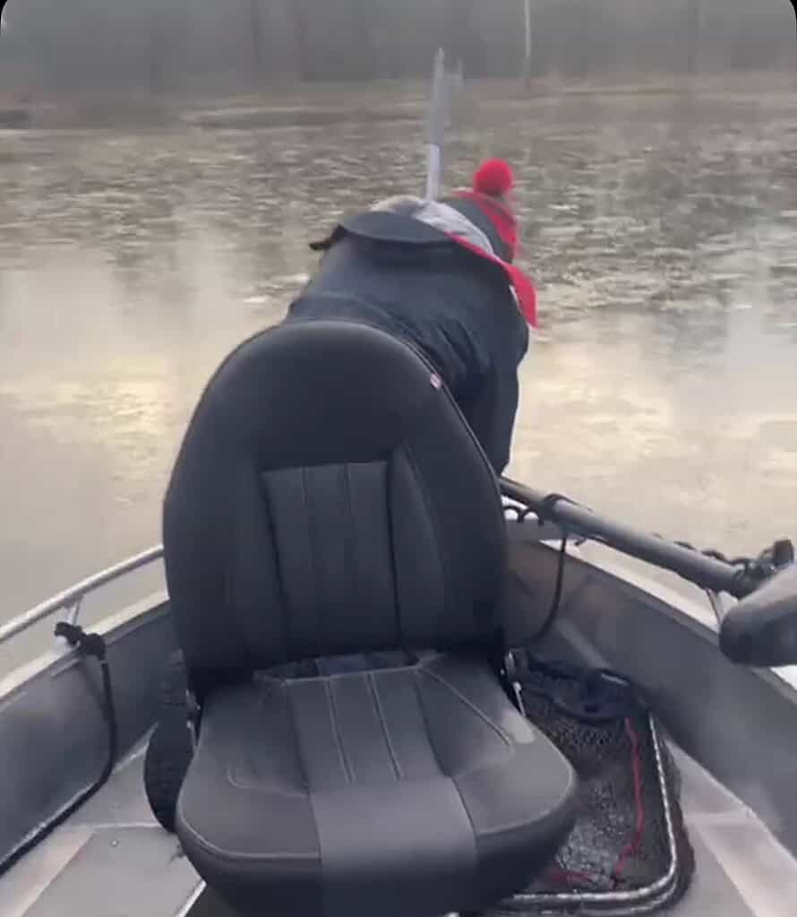 Een persoon met een rode hoed en jas reikt over de rand van een kleine boot op een kalm meer, misschien om te proberen doodaasvissen. Een lege bootstoel staat op de voorgrond.