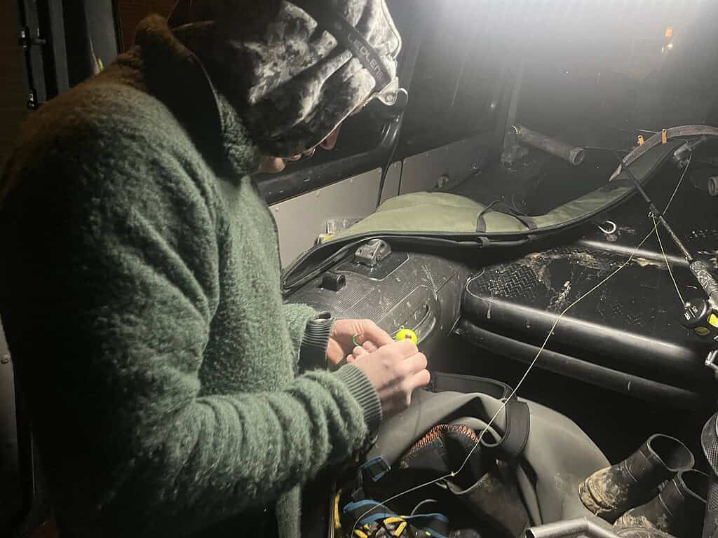 In een slecht verlicht voertuig knoopt een persoon in een groen jasje vakkundig knopen in vislijn, klaar om de strijd aan te gaan met de monsterlijke vis die onder het wateroppervlak op hem wacht.