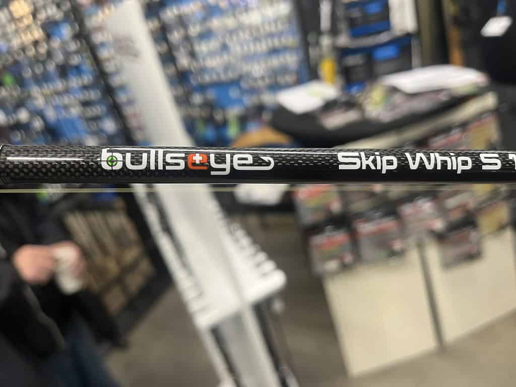 Close-up van een hengel met de tekst "Bullseye Skip Whip S", tentoongesteld in de winkel Hengelsport Maarssen, waar gezelligheid heerst tussen de wazige schappen en een persoon op de achtergrond.