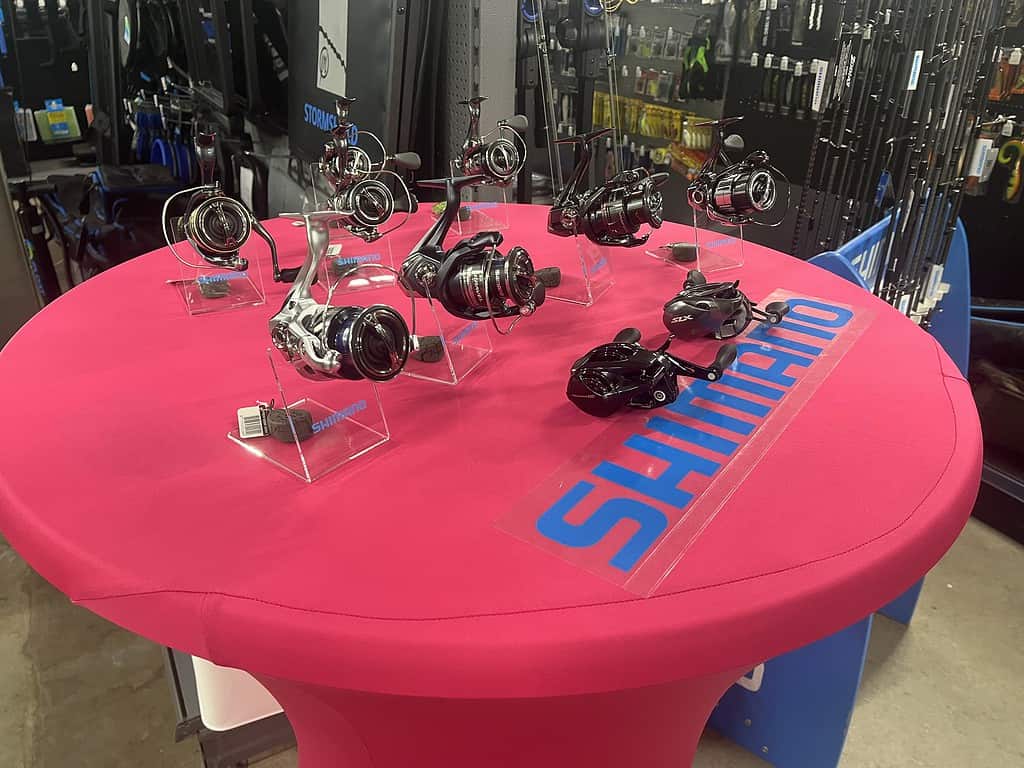 Op een ronde tafel met een rood tafelkleed met het Shimano-logo staan diverse vismolens uitgestald. Hierdoor ontstaat er een gezellige sfeer bij Hengelsport Maarssen, ideaal om de spullen te presenteren tijdens de Open Roofvis Dag.