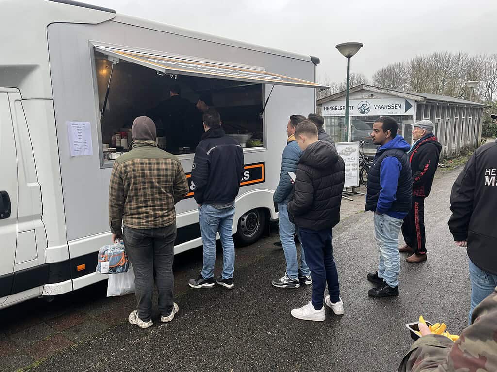 Mensen staan in de rij bij een foodtruck onder een bewolkte hemel tijdens de Open Roofvis Dag in Maarssen. Sommigen houden etenswaren vast, anderen wachten om te bestellen. Voldoende afstand tussen mensen suggereert een ongedwongen sfeer, perfect om hengelsporttips uit te wisselen terwijl je geniet van lekkernijen.