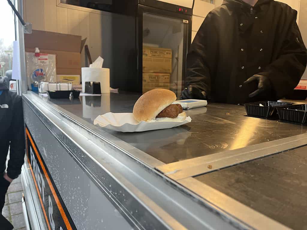 Een gepaneerde gehaktbal in een broodje op een wit servet staat op een metalen toonbank en straalt gezelligheid uit. Een persoon in donkere kleding staat ernaast in een foodtruck op het evenement Hengelsport Maarssen.