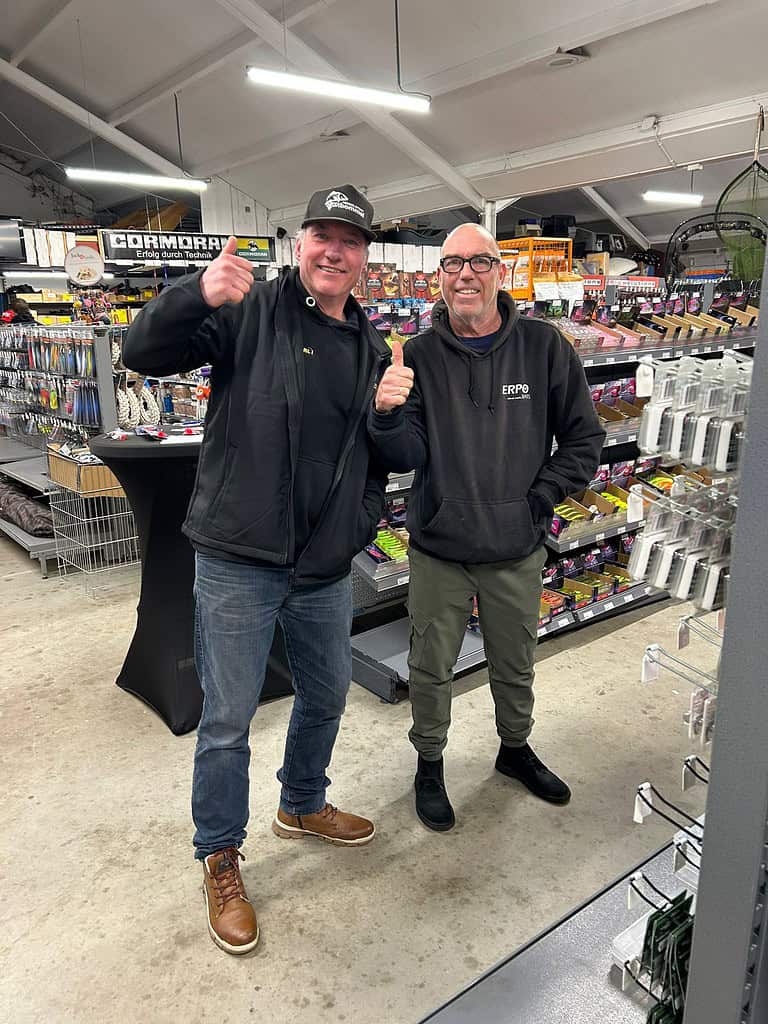 Twee mannen staan in een winkel in Maarssen, beiden met hun duim omhoog. Ze zijn casual gekleed en worden omringd door schappen vol hengelsportspullen, wat de geest van Open Roofvis Dag vastlegt.