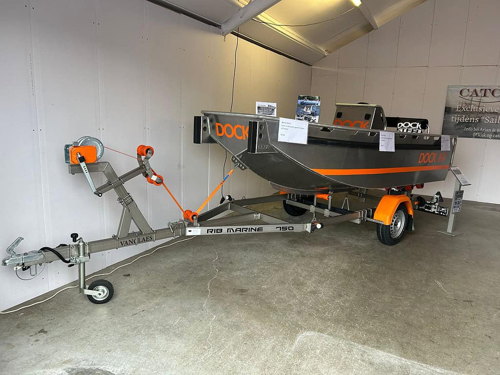 Een kleine grijs-oranje boot op een trailer staat in de garage, wachtend op de volgende Open Roofvis Dag. De trailer, gelabeld "RIQ MARINE 750," is uitgerust met een handlier en veiligheidsvoorzieningen, klaar om zowel avontuur als gezelligheid te bieden.