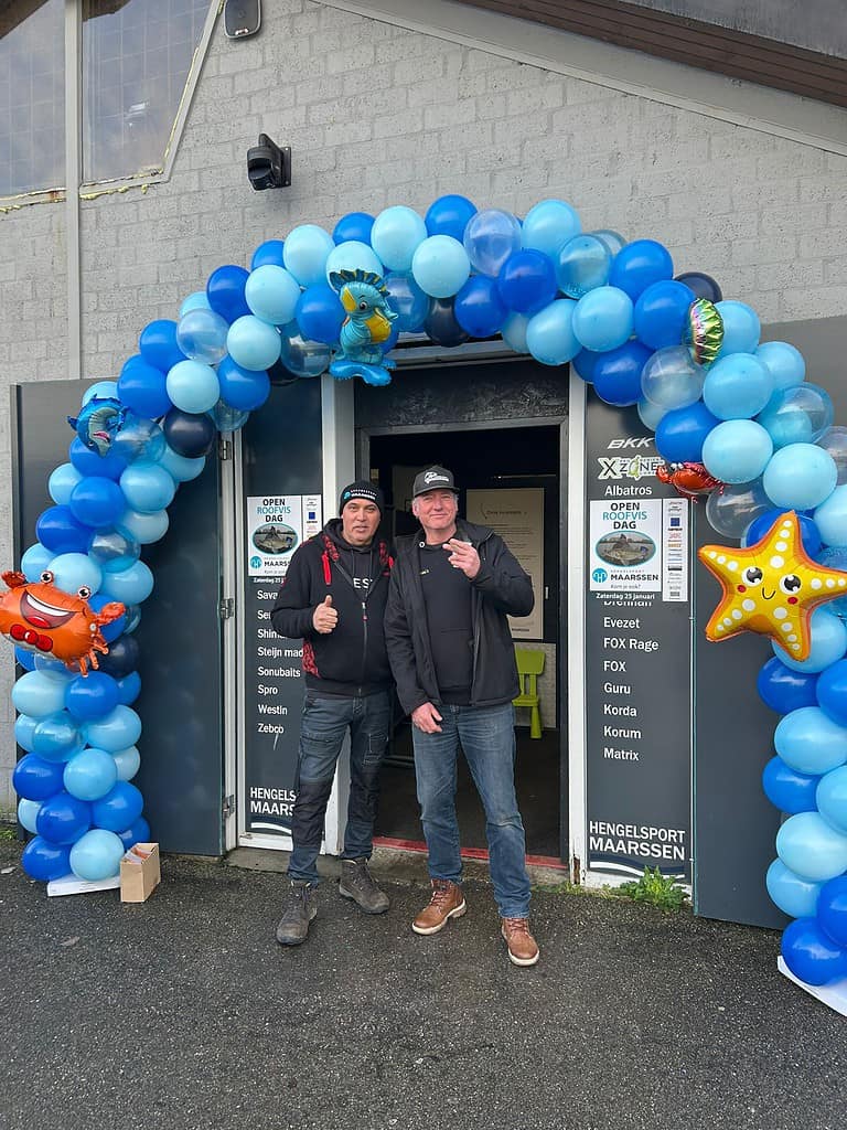 Twee mensen staan voor een deuropening versierd met blauwe en witte ballonnen en opblaasbare zee-thema's, die de gezelligheid van Open Roofvisdag belichamen. Beiden geven een duimpje omhoog, waarmee ze de geest van Hengelsport Maarssen vastleggen.