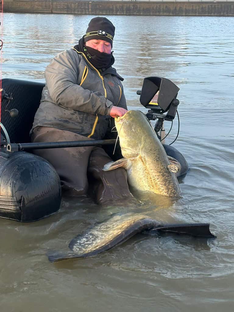 Een persoon in visgerei houdt trots een grote meerval vast terwijl hij op een opblaasboot in de plassen zit, met een betonnen muur op de achtergrond.