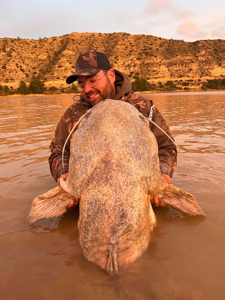 Een man in camouflagekleding houdt trots een grote vis vast terwijl hij in het water staat, tegen de achtergrond van het indrukwekkende berglandschap van de Ebro.