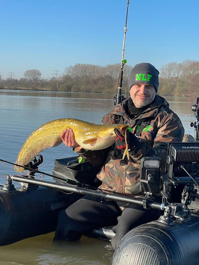 Een persoon in een boot houdt een grote vis vast, trots een camouflagejack en een "NLF"-muts dragend. Het serene water strekt zich uit tot aan de horizon onder de heldere hemel, waardoor het een perfecte dag is voor voorjaar vangsten.