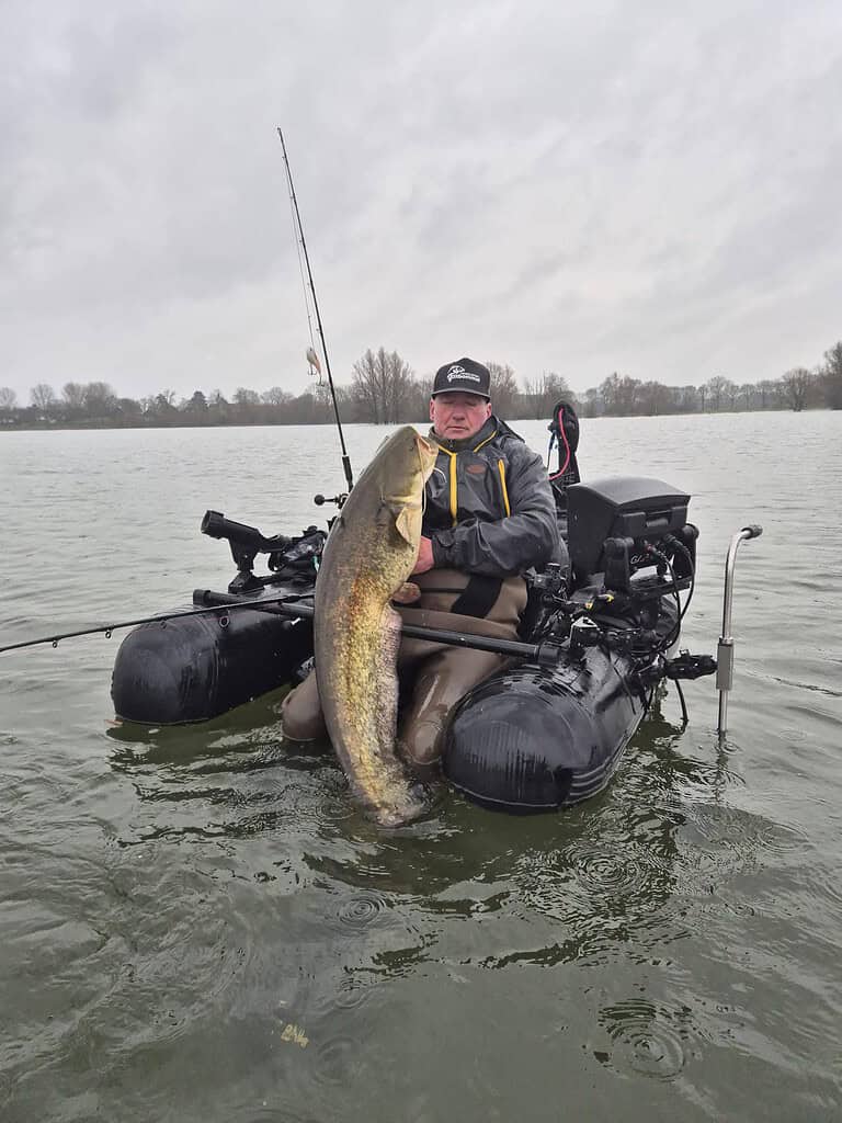 Een persoon toont trots een grote vangsten in zijn boot, omringd door visgerei op een kalm meer. De bewolkte lucht strekt zich uit over de horizon en vat de rust van een kalme voorjaarsdag samen.