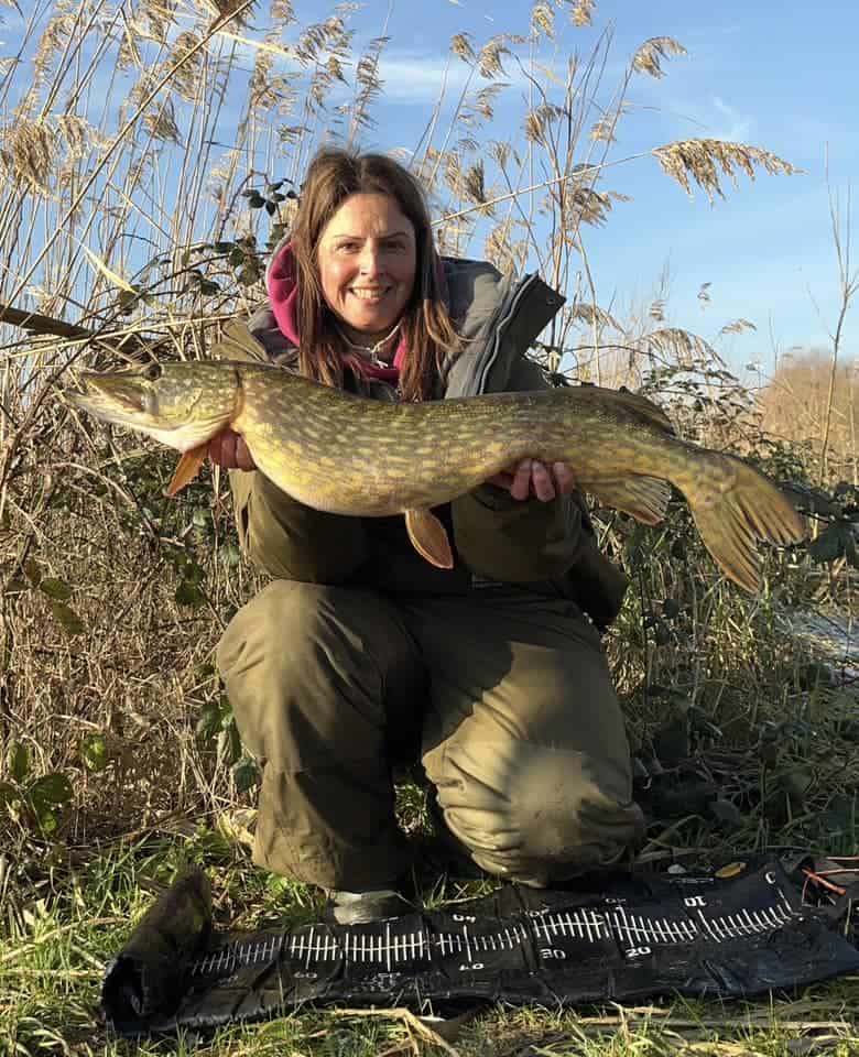 Persoon knielt buiten, met een grote vis met groene en gele patronen, gevangen tijdens een vruchtbare voorjaar. Omringd door hoog gras en riet, omlijst de uitgestrekte horizon het tafereel, terwijl een meetmat gedeeltelijk zichtbaar is op de grond.