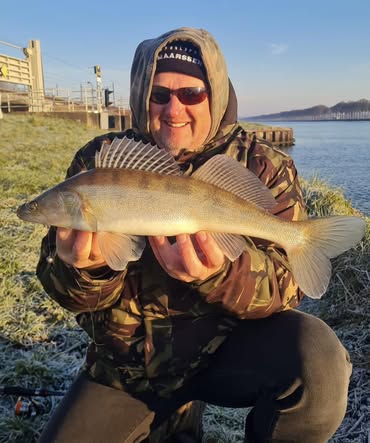 Persoon in een camouflagejack met een vis in zijn handen aan de oever van de rivier, glimlachend onder een heldere hemel en met de horizon die zich eindeloos uitstrekt.