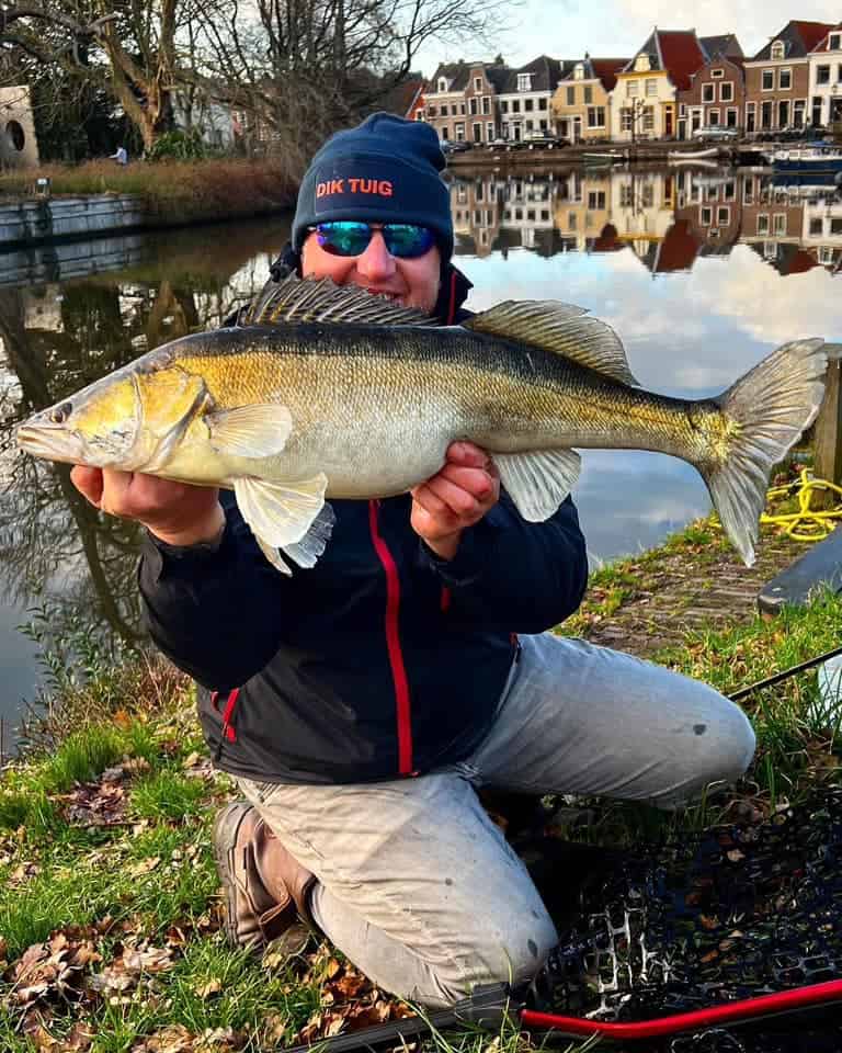 Een persoon knielt bij een kanaal en toont trots een van de vangsten van de week, een grote vis. Op de achtergrond staan charmante huizen aan de waterkant, wat een pittoreske setting creëert.