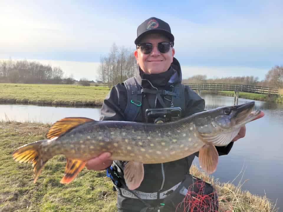 Een persoon toont trots een grote vis aan de rivier, met een pet, zonnebril en Predatorgear-visserskleding. Op de achtergrond strekt een brug zich uit over de grazige oever onder de heldere hemel - een perfecte dag voor Vangsten van de maand.