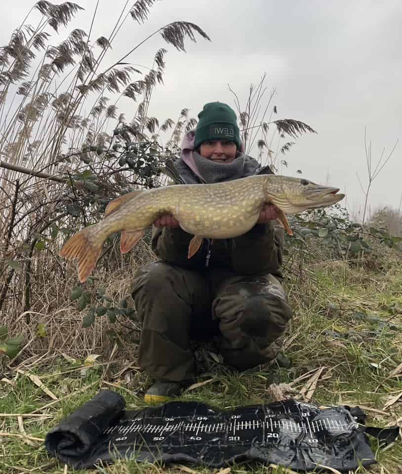 Een trotse visser in Predatorgear poseert met een grote vis bij het riet. Het is de vangst van de maand en hij staat tentoongesteld op een meetmat eronder.