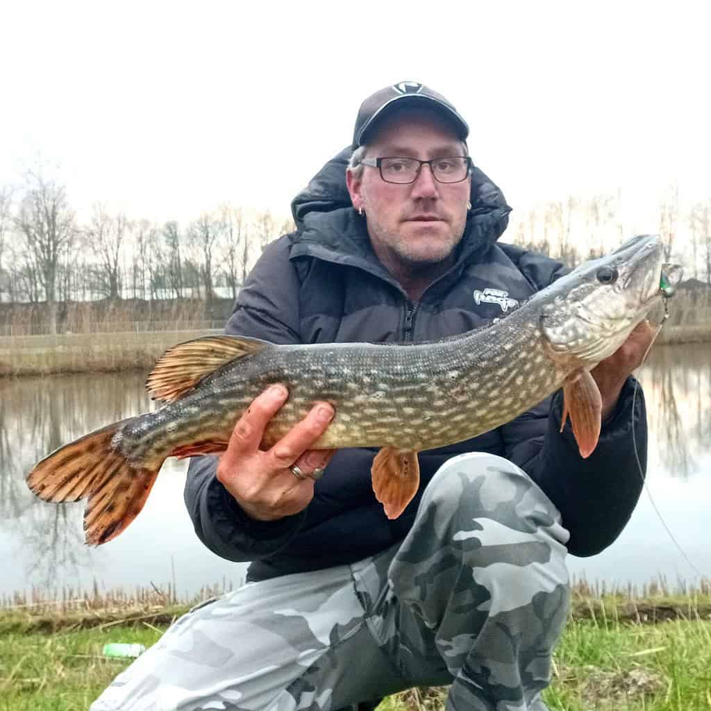 Een persoon die trots zijn vangsten laat zien aan de oever van het meer, gekleed in een zwart jasje en camouflagebroek. De serene achtergrond wordt omlijst door bomen en glinsterend water, wat de perfecte week van de schoonheid van de natuur vastlegt.