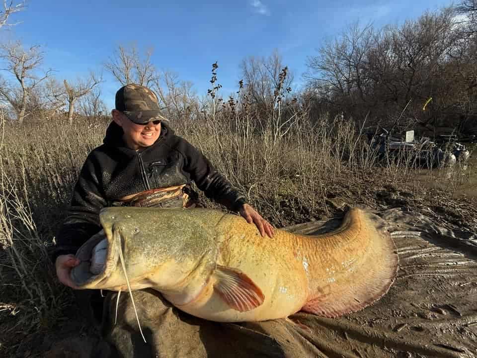 Met Predatorgear aan zit de persoon op de modderige grond, trots een grote meerval vasthoudend. Spaarzame bomen en droge vegetatie vormen de achtergrond onder een helderblauwe lucht. Het is een perfect vangsten-moment, vastgelegd in de serene omhelzing van de natuur.