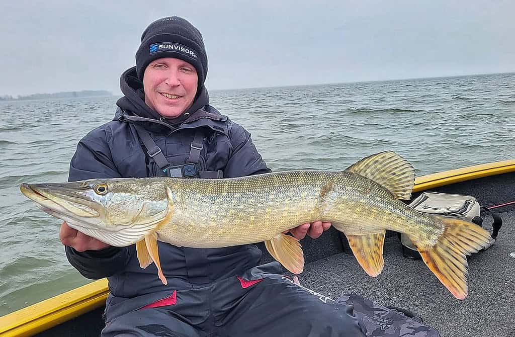 Persoon op een boot die een grote vis vasthoudt tegen een achtergrond van een bewolkte lucht en water, die trots de vangsten laat zien. Gekleed in Predatorgear's zwarte jas en hoed, belichamen ze de sensatie van de vangst.