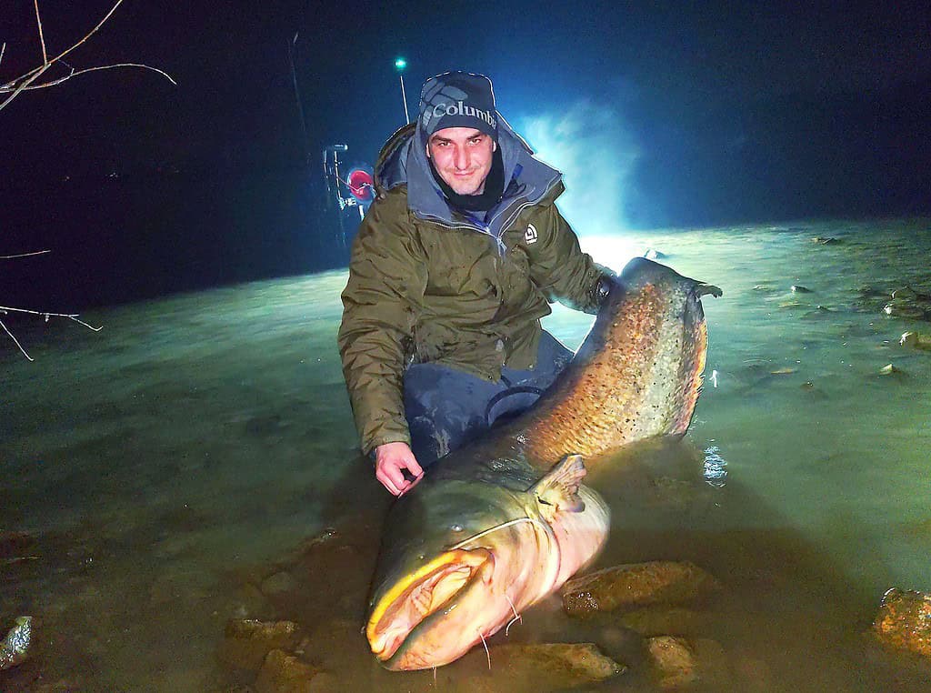 's Nachts knielt iemand in ondiep water, gekleed in een winterjas en een hoed, en toont trots zijn nieuwste vangsten, een grote vis.