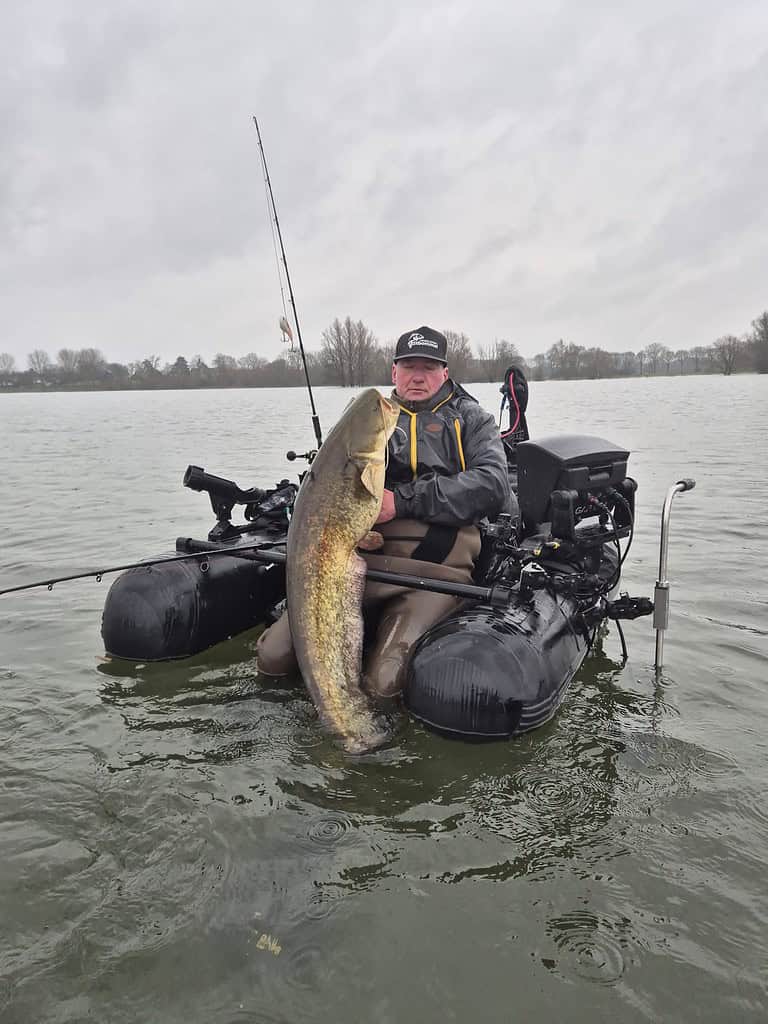 Een persoon zit op een kleine opblaasbare pontonboot en houdt trots een grote vis vast die hij tijdens de wintermaanden heeft gevangen. Hengels rusten in de buurt onder de bewolkte lucht, wat een succesvol vissenavontuur laat zien.