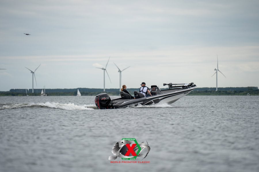 Een motorboot vaart over een meer met twee mensen aan boord. Windturbines en een zeilboot in de verte zijn zichtbaar op de achtergrond.